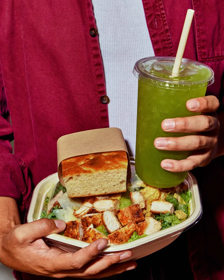 A sweetgreen Ginger Greens housemade beverage held alongside a warm bowl and a square of focaccia