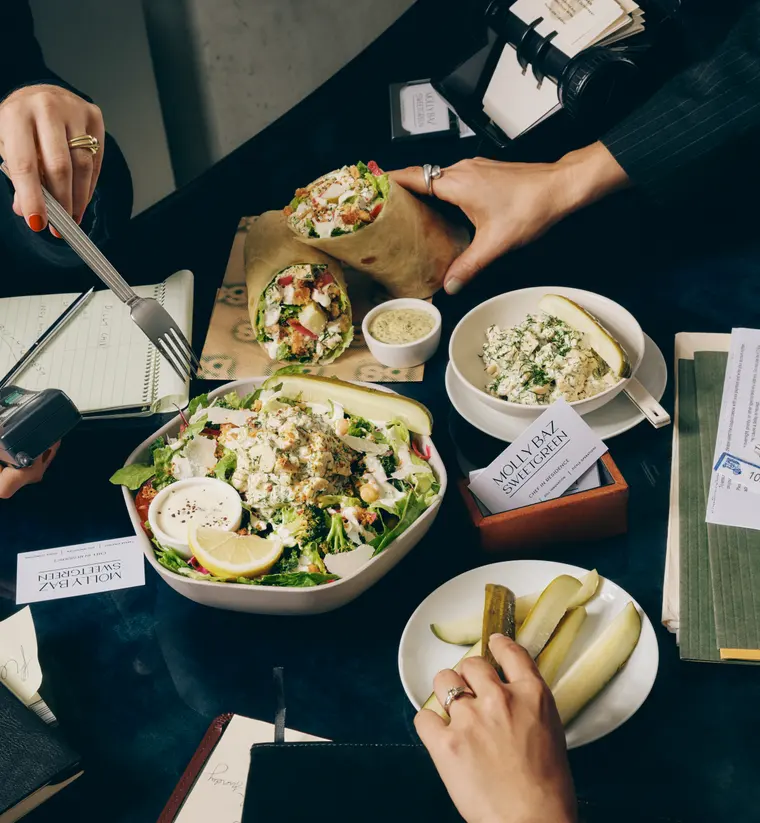 An overhead desk spread of Molly's wraps, Pickle Caesar Sal, chicken salad scoop and pickle spears shared between two people