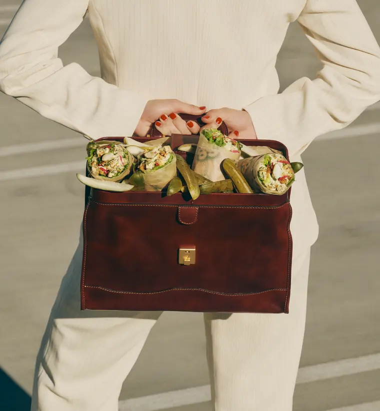 Molly Baz holding an open leather briefcase packed with her sweetgreen wraps and pickle spears