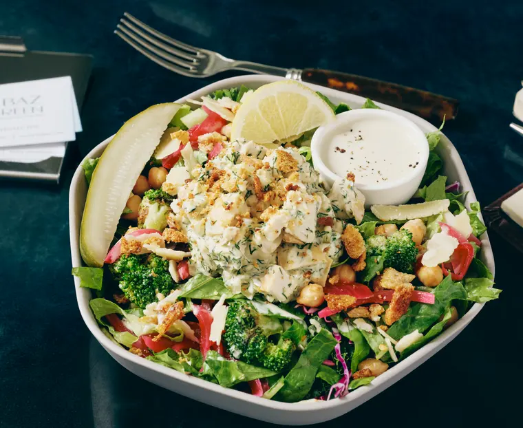 Molly's Pickle Caesar Sal, a pickle Caesar salad in a white bowl with chicken salad, spicy broccoli, garlic breadcrumbs, a lemon wedge and a whole pickle spear