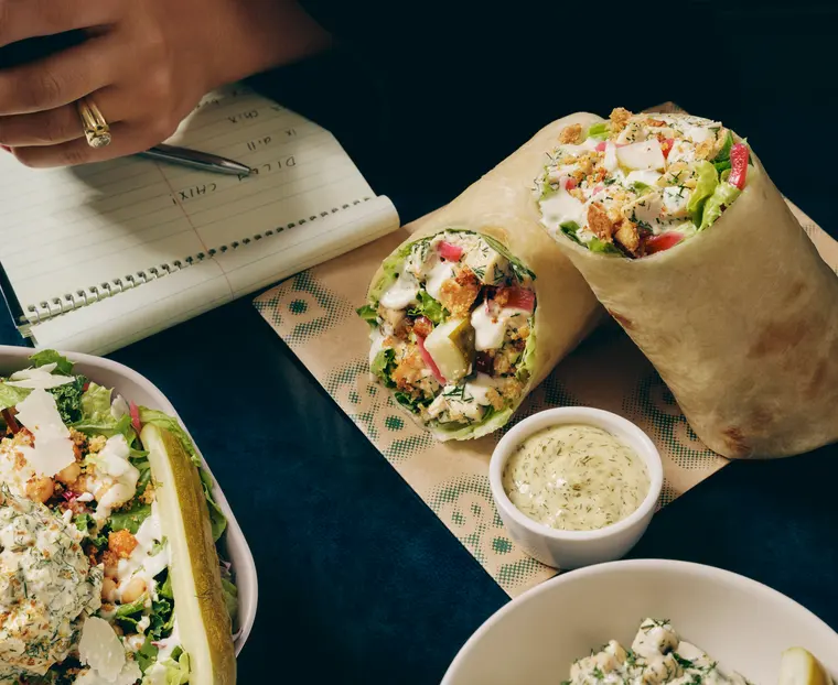 Molly's Big Dill Caesar Wrap cut in half, showing chicken salad, romaine, fresh dill and pickled red onion in an olive oil tortilla, with a cup of Caesar dressing alongside