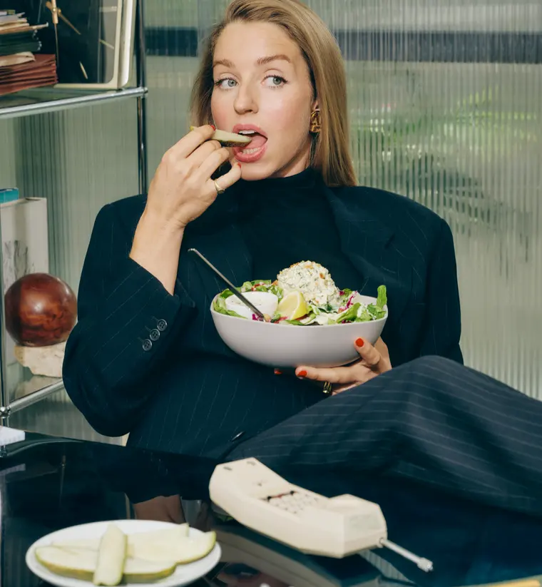 Molly Baz at a desk eating a pickle slice, holding a bowl of her Pickle Caesar Sal